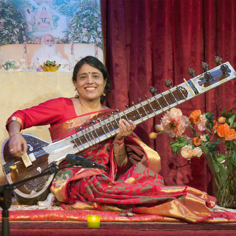 A woman in a red sari plays a sitar in the foreground, while a tapestry depicting a spiritual figure hangs in the background.