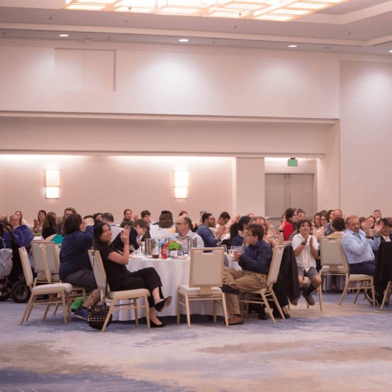 Guests are seated at round tables in a brightly lit banquet hall with a patterned carpet in the foreground and a stage area in the background.