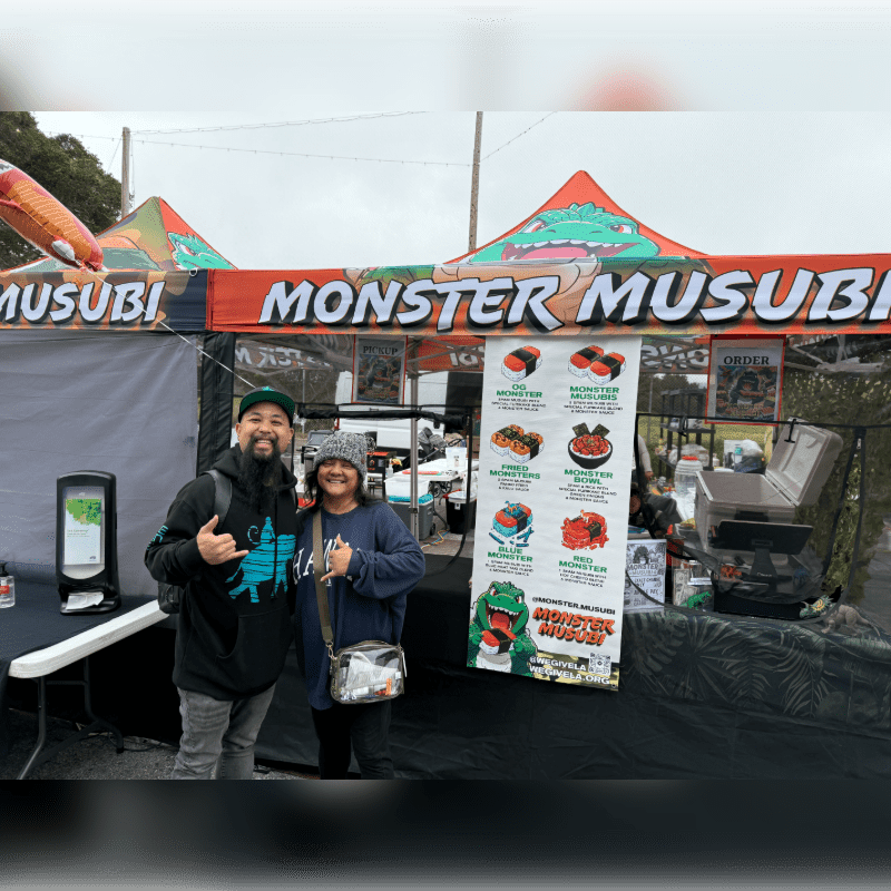 A couple poses in front of a "Monster Musubi" food tent with a menu board and a cooler in the background.