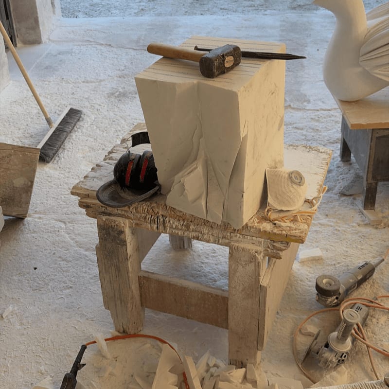 A rough stone block sits on a wooden stool in the foreground, with sculpting tools and debris scattered around, while a partially carved white bird sculpture is visible in the background.