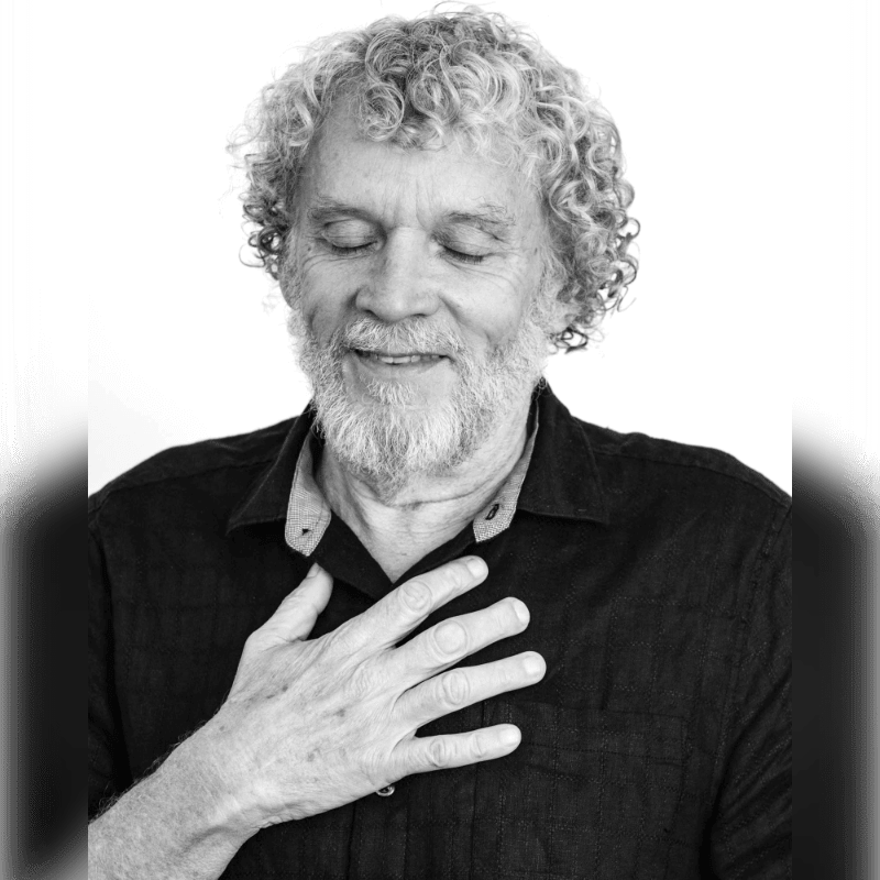 A man with curly gray hair and a beard places his hand over his heart with his eyes closed against a plain white background.