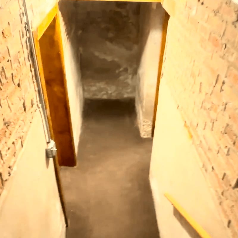 A dimly lit basement hallway with exposed brick walls in the foreground leads to a dark, unfinished space in the background.