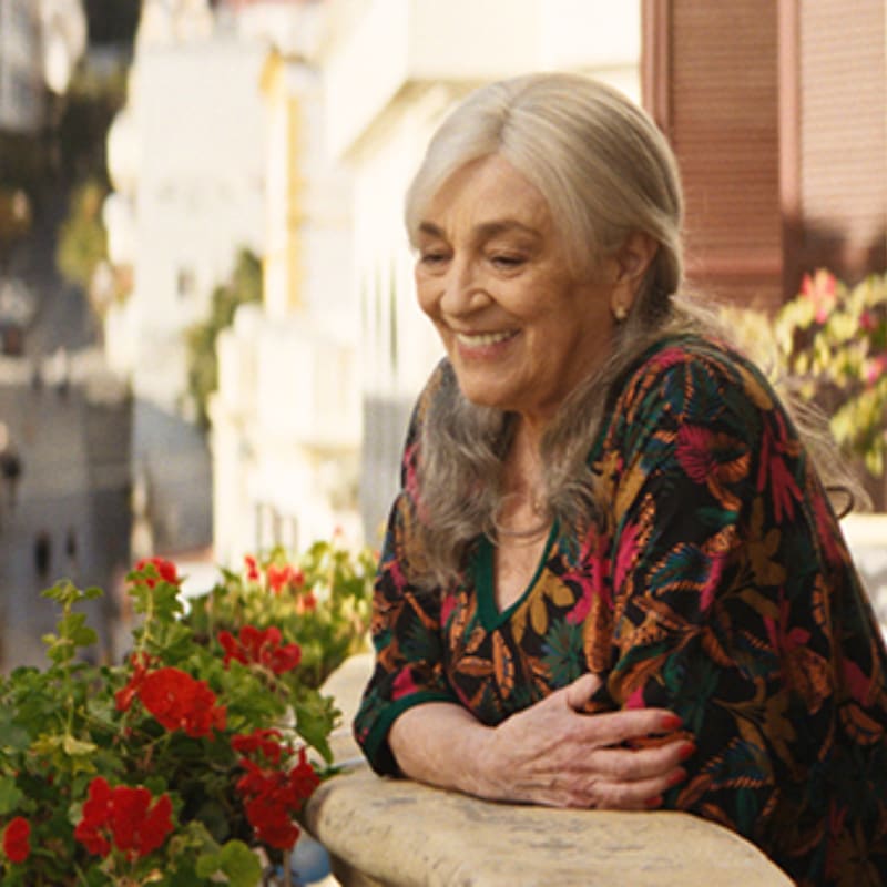 A smiling elderly woman with gray hair leans on a balcony railing with red flowers in the foreground, while a blurred cityscape is visible in the background.