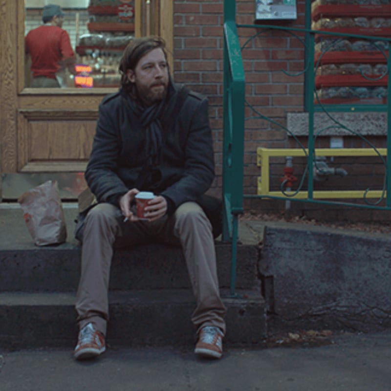 A man with a beard sits on concrete steps holding a coffee cup, with a brick building and a store window in the background.
