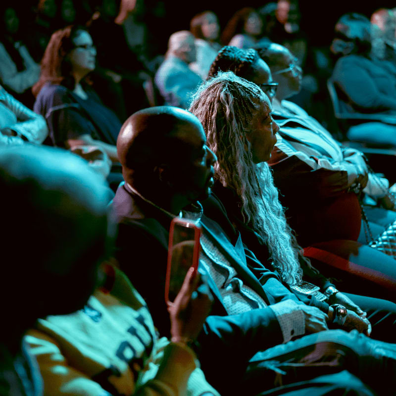 In the foreground, a person holds up a red smartphone, while the background is filled with a dimly lit audience watching an event.