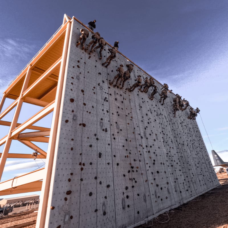 Soldiers climb a wooden obstacle wall with a clear blue sky and a distant airplane in the background.