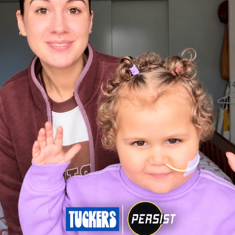 A woman smiles behind a young child with curly hair and a feeding tube, both in casual clothing, with a blurred background of a room.