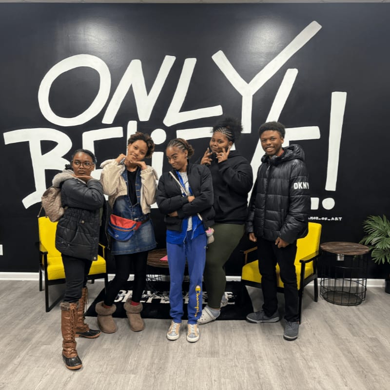 Five young people pose in front of a large black wall with the words "ONLY BELIEVE!" painted in white, with yellow chairs flanking them.