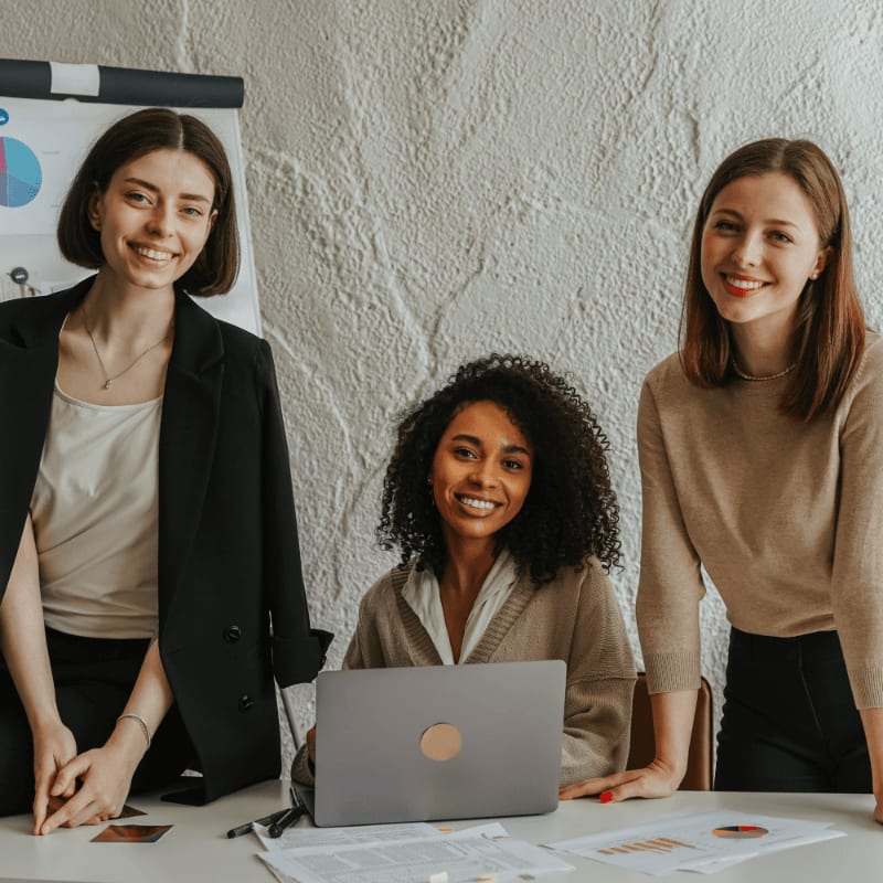 Three smiling women are gathered around a table with a laptop and papers, with a whiteboard in the background.