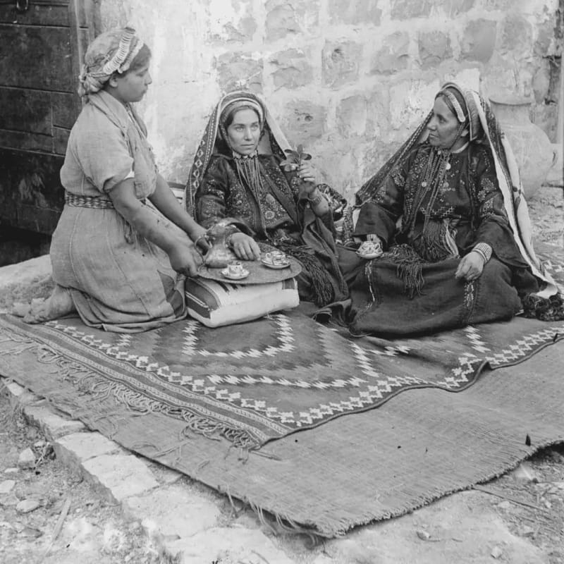 Three women in traditional attire sit on a patterned rug in the foreground, with a stone wall and wooden door forming the background.