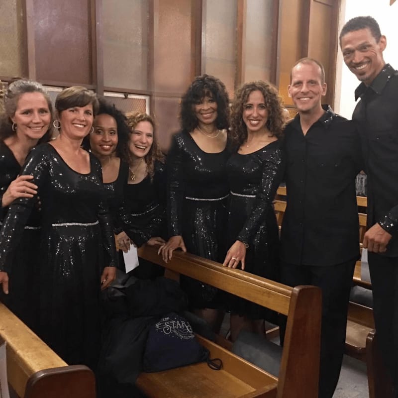 A group of seven singers in sparkling black dresses and shirts stand in front of wooden pews and a wall with vertical panels.