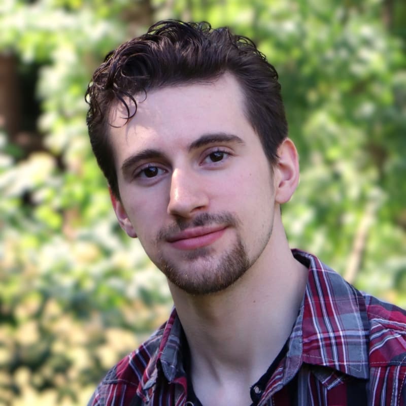 A young man with dark hair and a goatee, wearing a plaid shirt, smiles gently against a blurred background of green foliage.
