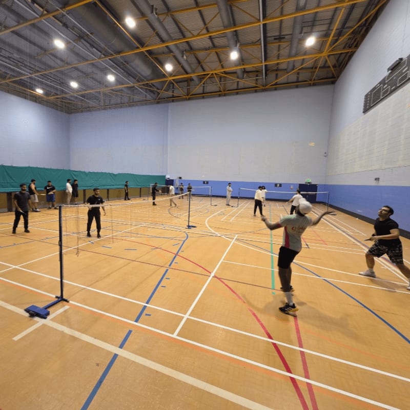 People play badminton on a wooden court with nets set up, while others watch from the sidelines in the background of a brightly lit indoor gymnasium.