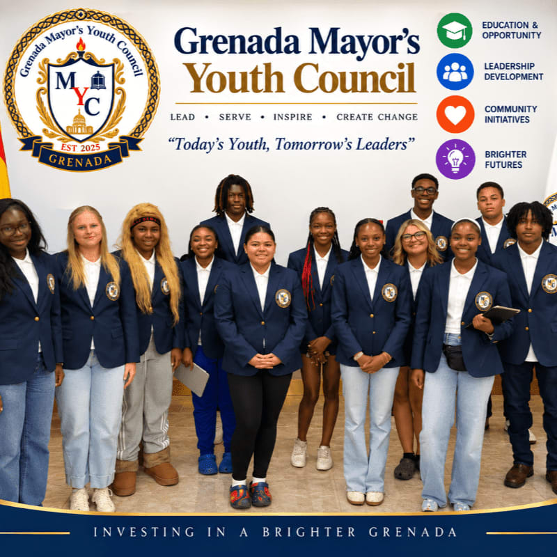A group of young people in matching blazers stand in front of a wall displaying the Grenada Mayor's Youth Council logo and mission statement.