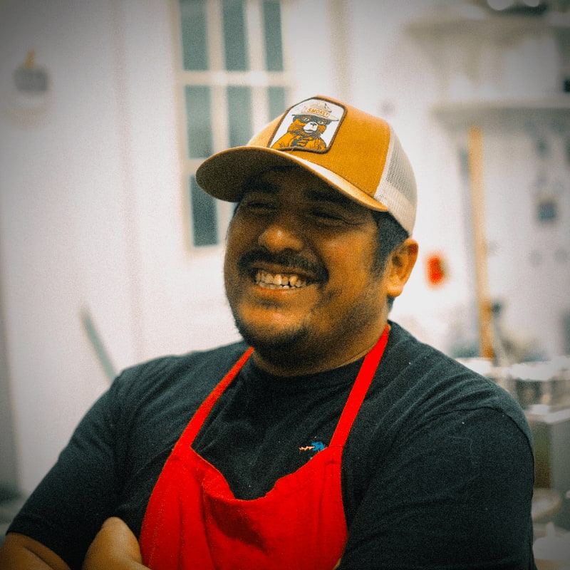 A man in a red apron and a trucker hat smiles in the foreground, with a blurred kitchen in the background.
