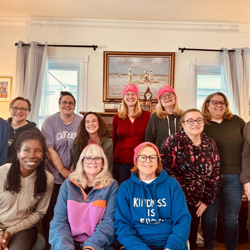 A group of women wearing pink beanies pose for a photo in a room with a fireplace and artwork.