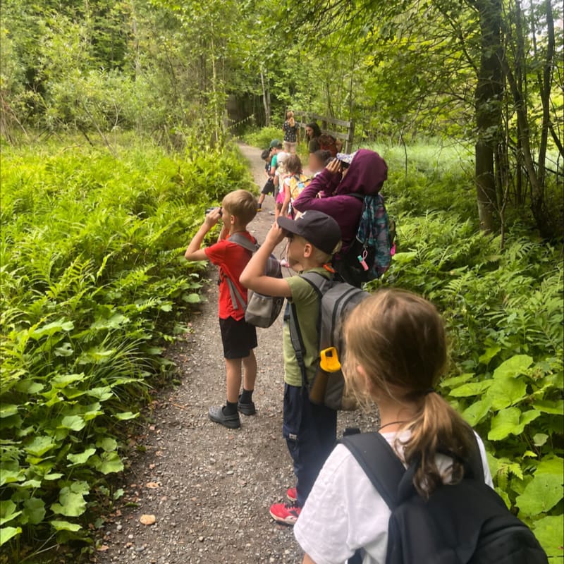 Children in the foreground look through binoculars on a path surrounded by lush green foliage, while a group of people walks further down the trail in the background.