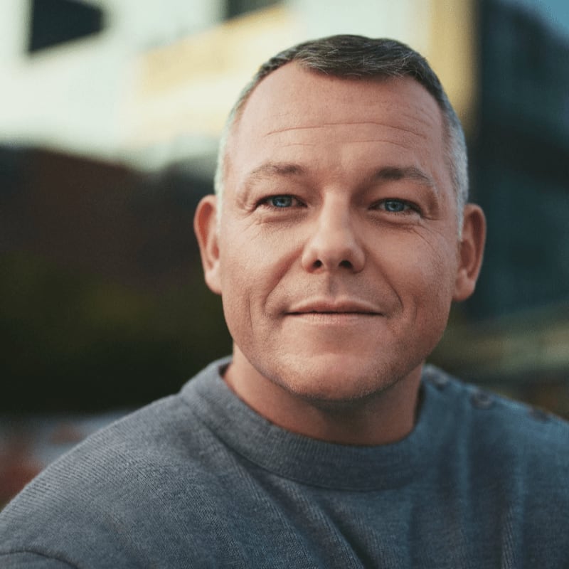 A man with short gray hair and blue eyes smiles gently in the foreground, with a blurred urban background behind him.