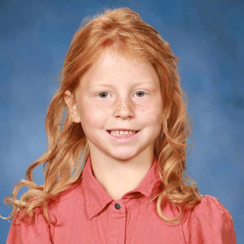 A young girl with red hair and freckles smiles in the foreground against a blue background.