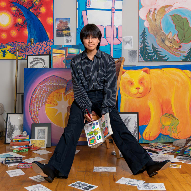 A person sits cross-legged on a chair holding a sketchbook, surrounded by colorful paintings and art supplies scattered on a wooden floor.