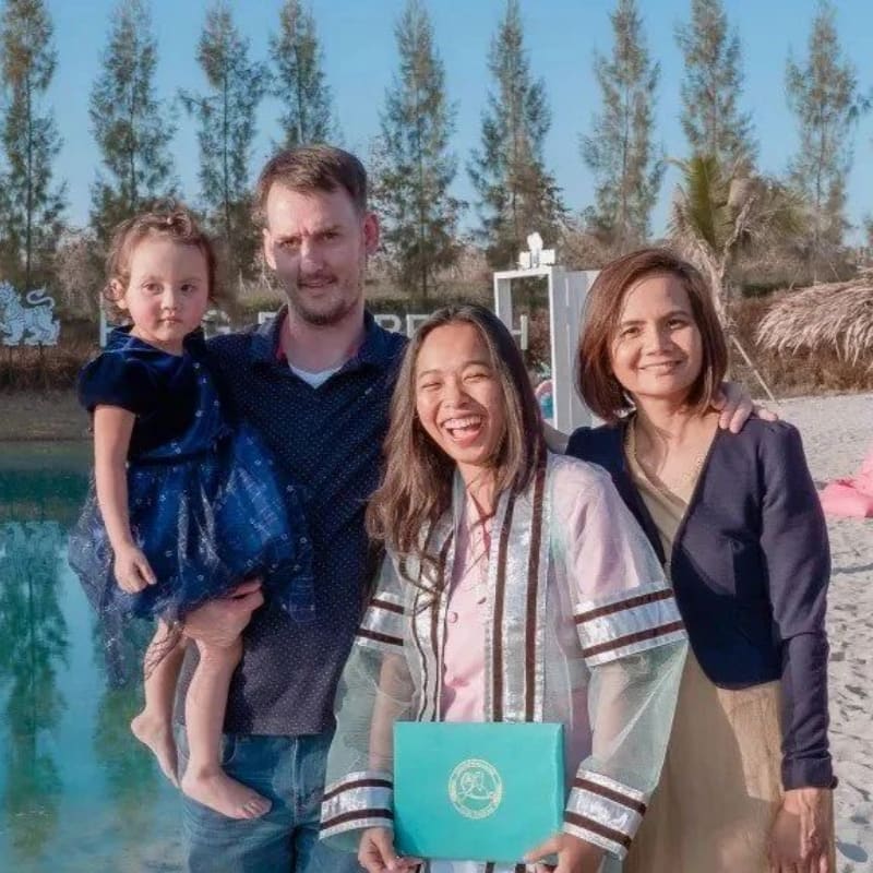 A family poses for a photo on a sandy beach with a body of water and trees in the background.