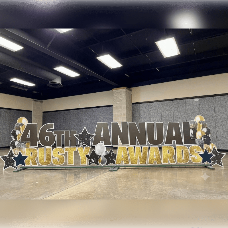 Large, glittery letters spelling "46TH ANNUAL RUSTY AWARDS" stand in the foreground, adorned with balloons and stars, against a backdrop of a large, empty room with a tiled floor and acoustic paneling on the walls.