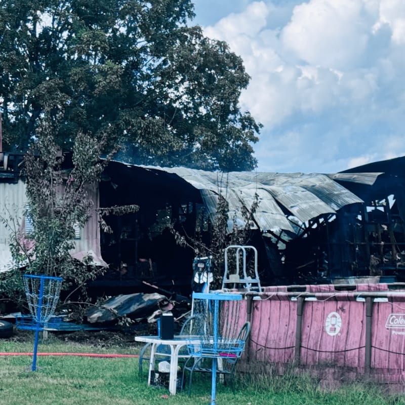 A partially burned trailer home with a collapsed roof stands in the background, while a pink above-ground pool and a small table with chairs are in the foreground.