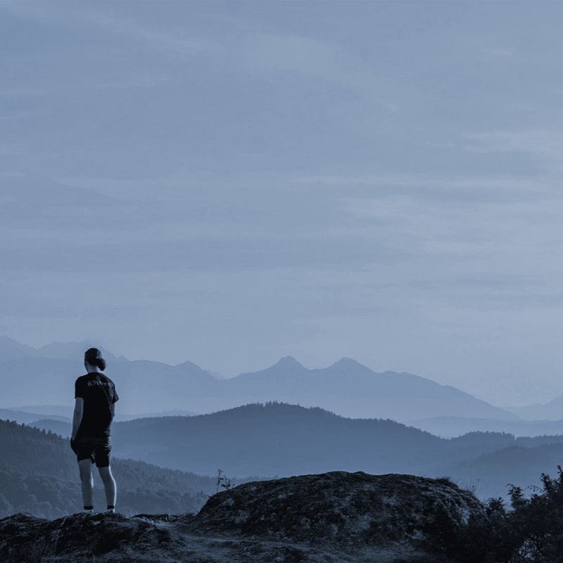 A lone figure stands on a rocky outcrop, gazing out at a vast expanse of misty blue mountains.