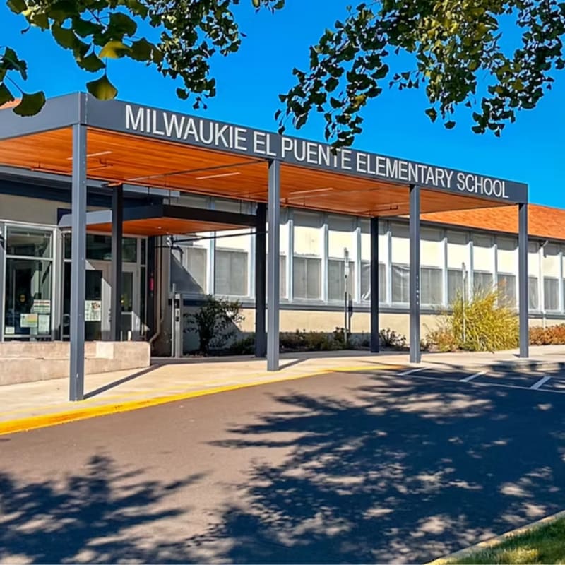 A paved walkway leads to the entrance of Milwaukie El Puente Elementary School, with a clear blue sky and tree branches overhead.