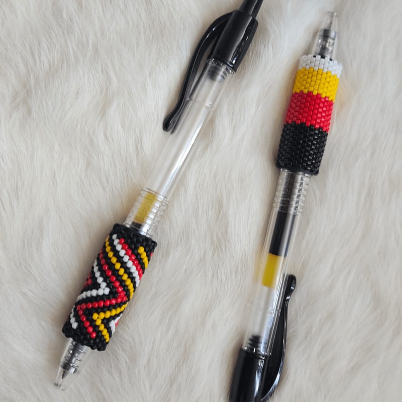 Two pens with beaded grips in red, yellow, and black patterns rest on a white, fluffy surface.