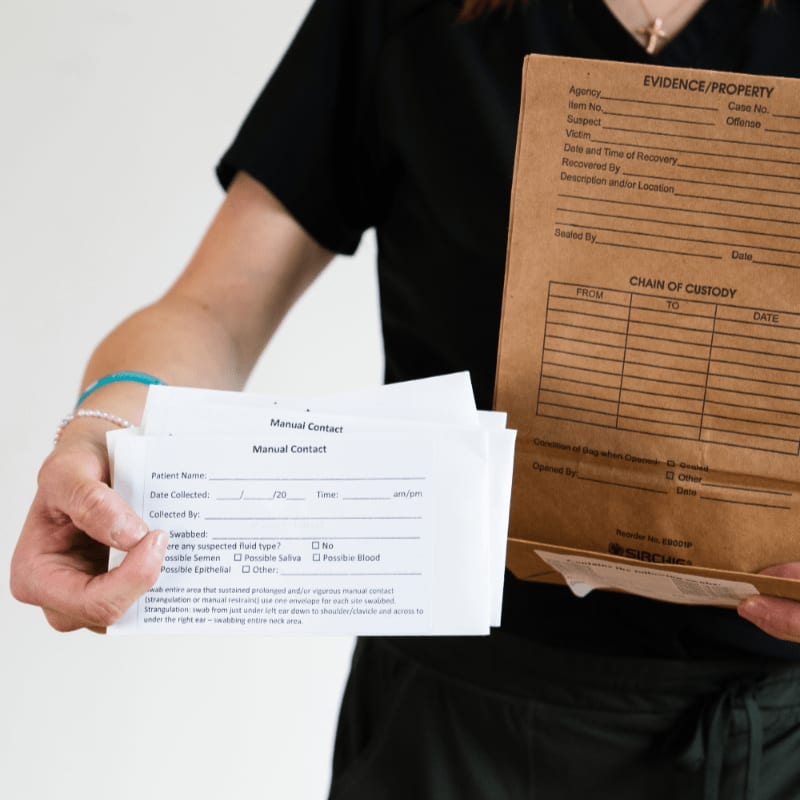 A person in dark scrubs holds a brown evidence bag and several white forms in front of a white background.