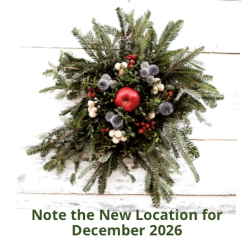 A festive evergreen wreath with a red apple, berries, and thistles is centered against a white wooden plank background, with text below stating "Note the New Location for December 2026".