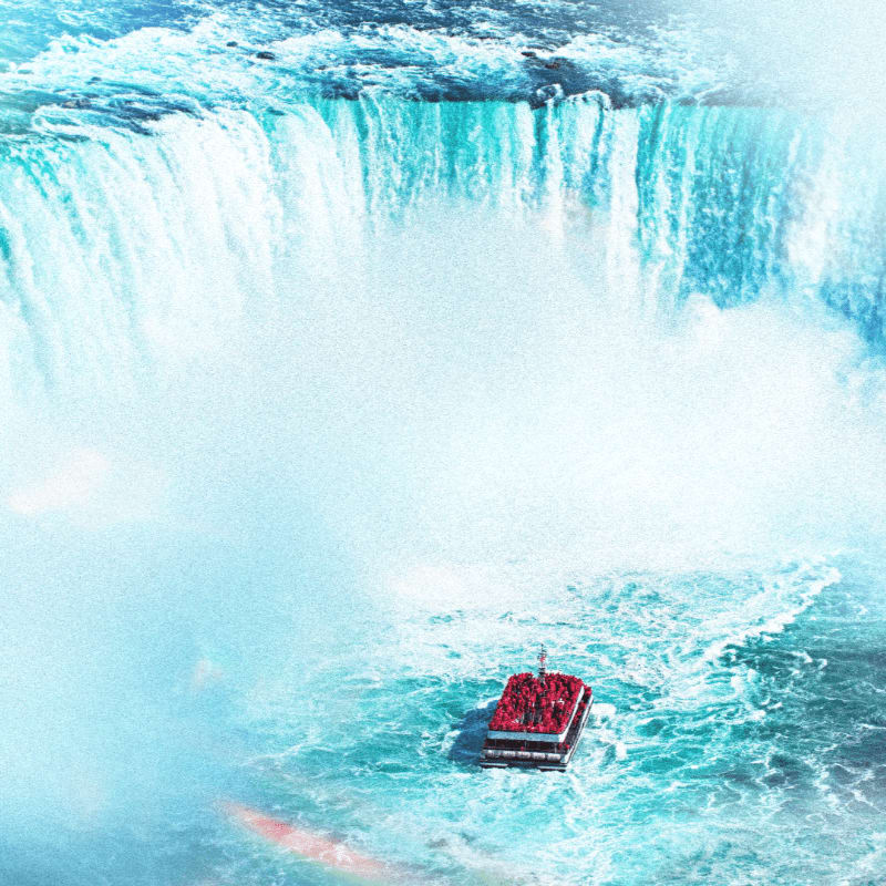 A red boat with people on its deck navigates the turbulent, mist-shrouded waters at the base of a massive, cascading waterfall.