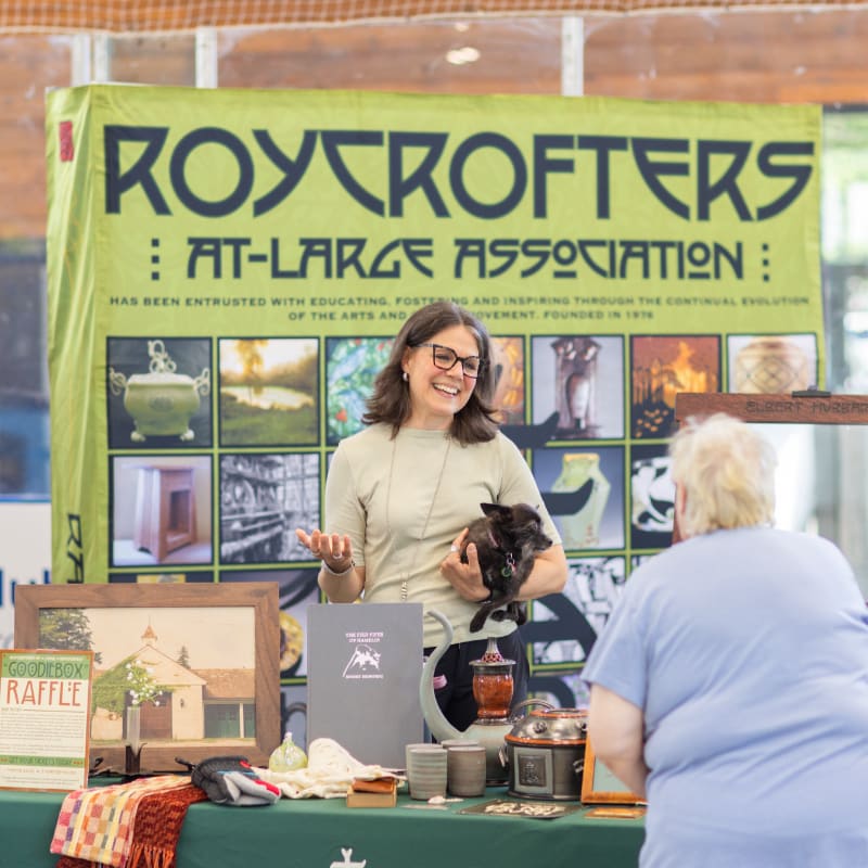 A woman with a dog stands behind a table of crafts in front of a Roycrofters banner.