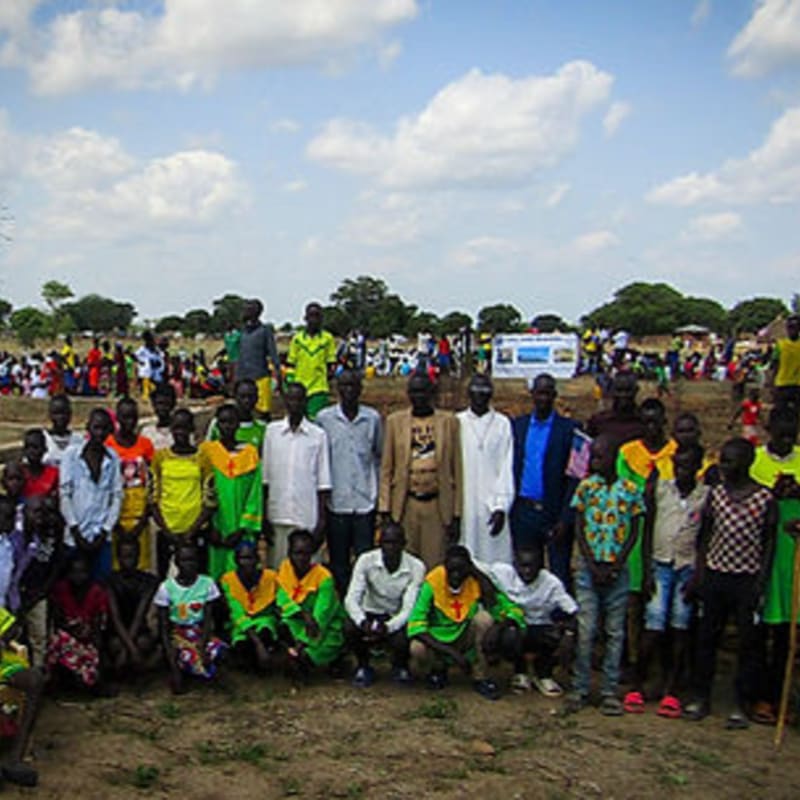 A group of people, many in brightly colored shirts, pose for a photo in the foreground with a larger crowd and trees in the background under a cloudy sky.