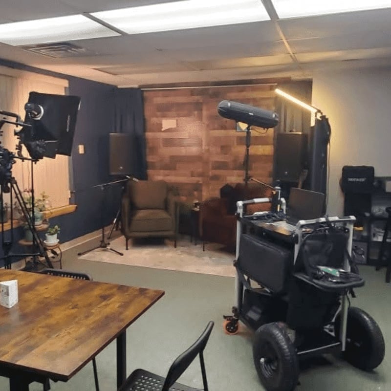 In the foreground, a wooden table and chairs are set up, while the background features a small studio with seating, lighting equipment, and a wood-paneled wall.