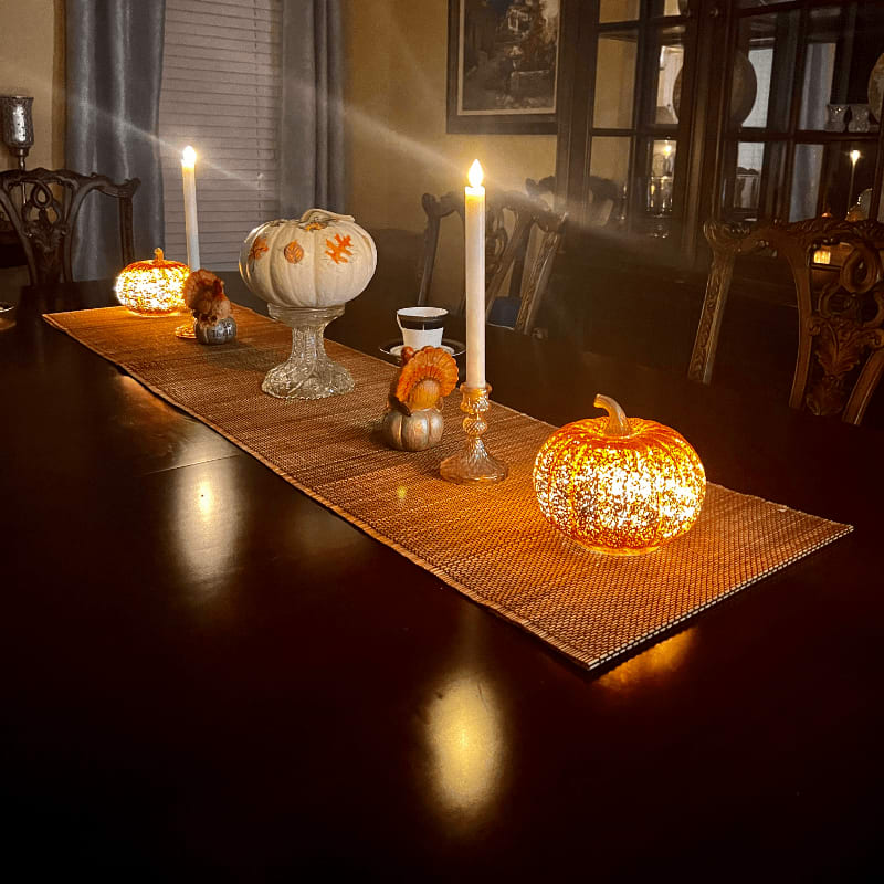 Illuminated pumpkins and candles adorn a table runner on a dark wood table, with a dimly lit dining room and ornate furniture in the background.