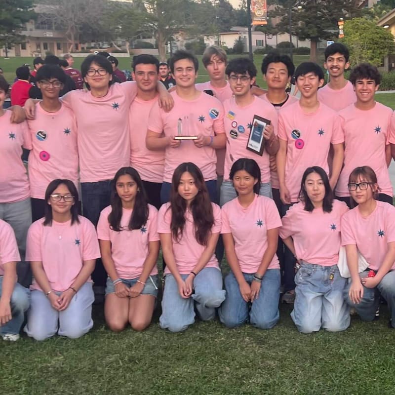 A group of young people in matching pink shirts pose for a photo on a grassy lawn with buildings and trees in the background.
