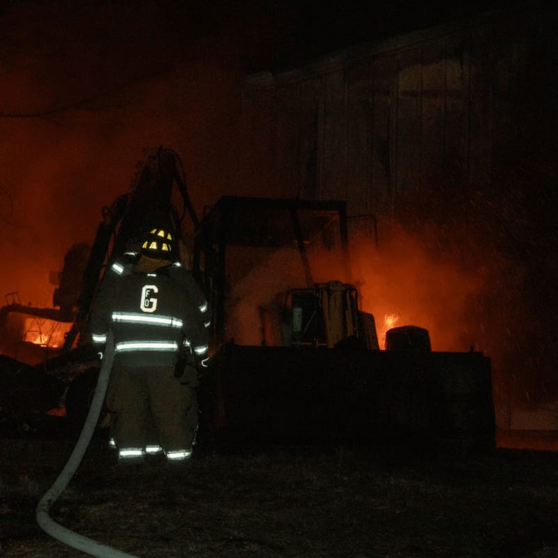 A firefighter in protective gear stands in the foreground holding a hose, while a large piece of machinery burns intensely in the background.