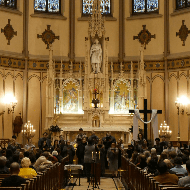A choir sings in the foreground of a church with a large cross and altar in the background.