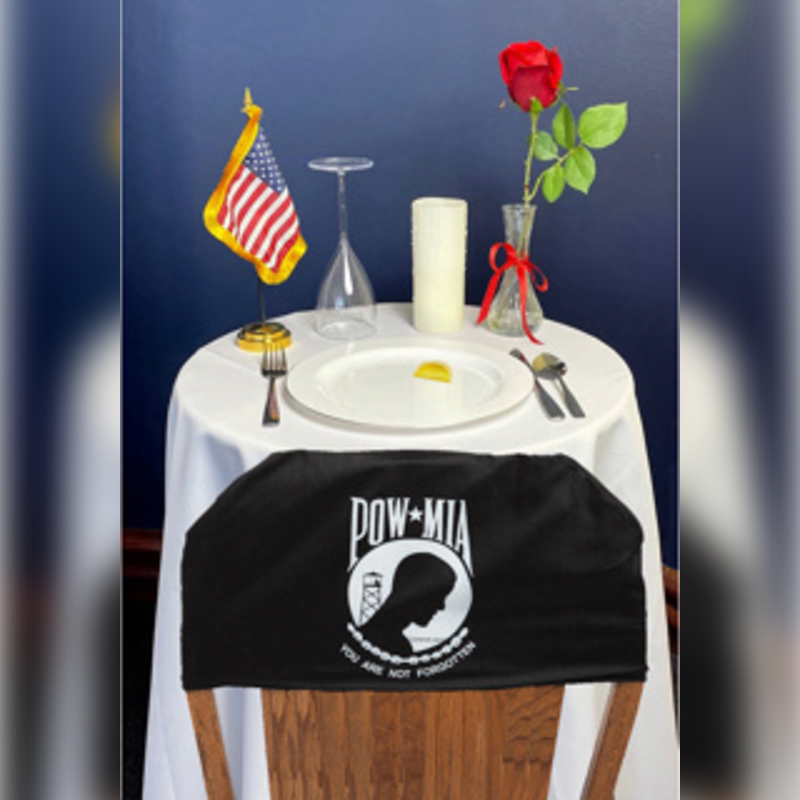 A single place setting with a POW-MIA flag draped over the chair sits on a table adorned with a rose and candle, against a dark blue background.