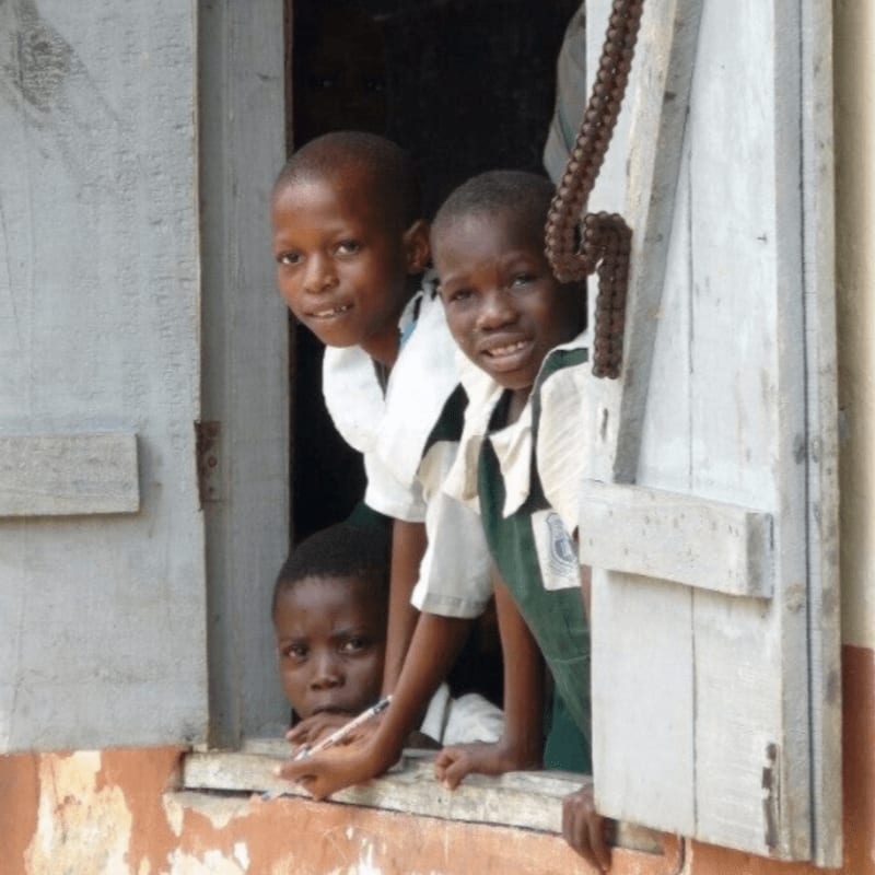 Three young children in school uniforms peer out of a weathered window frame, with a dark interior visible behind them.