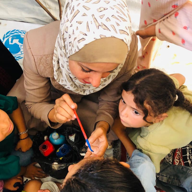 A woman in a hijab paints a child's face with bright colors, while other children watch intently in the background, with a UN logo visible on a tent.