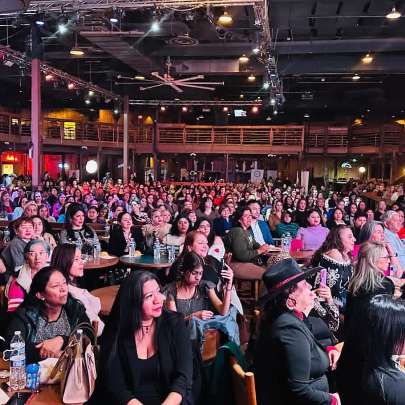 A large audience fills a dimly lit venue with wooden balconies and stage lighting in the background, while several women in the foreground are seated at tables.