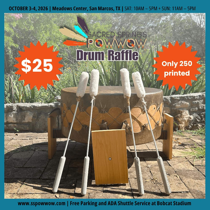 Four drumsticks rest against a large drum in the foreground, with a stone wall and greenery in the background.