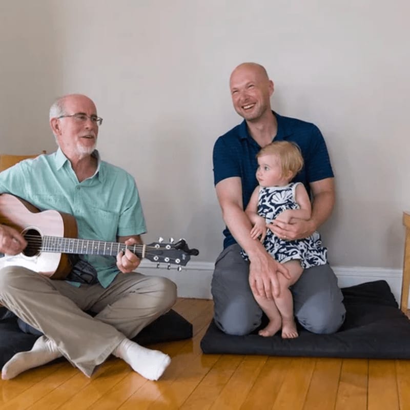 An older man plays guitar while a younger man holds a baby, with a plain wall and wooden floor in the background.