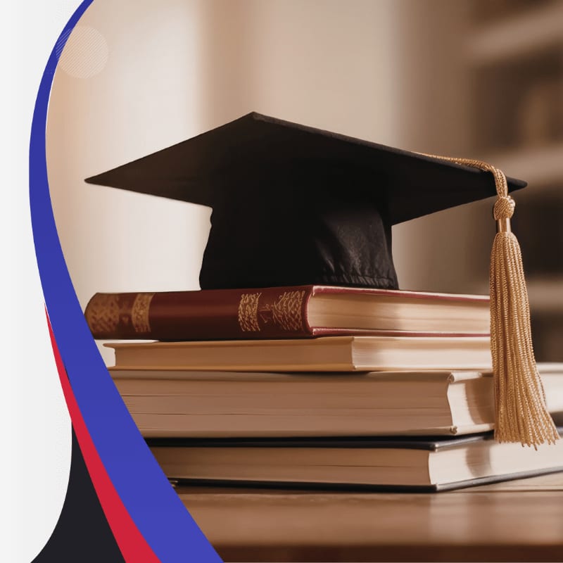 A graduation cap rests atop a stack of books in the foreground, with a blurred bookshelf in the background.