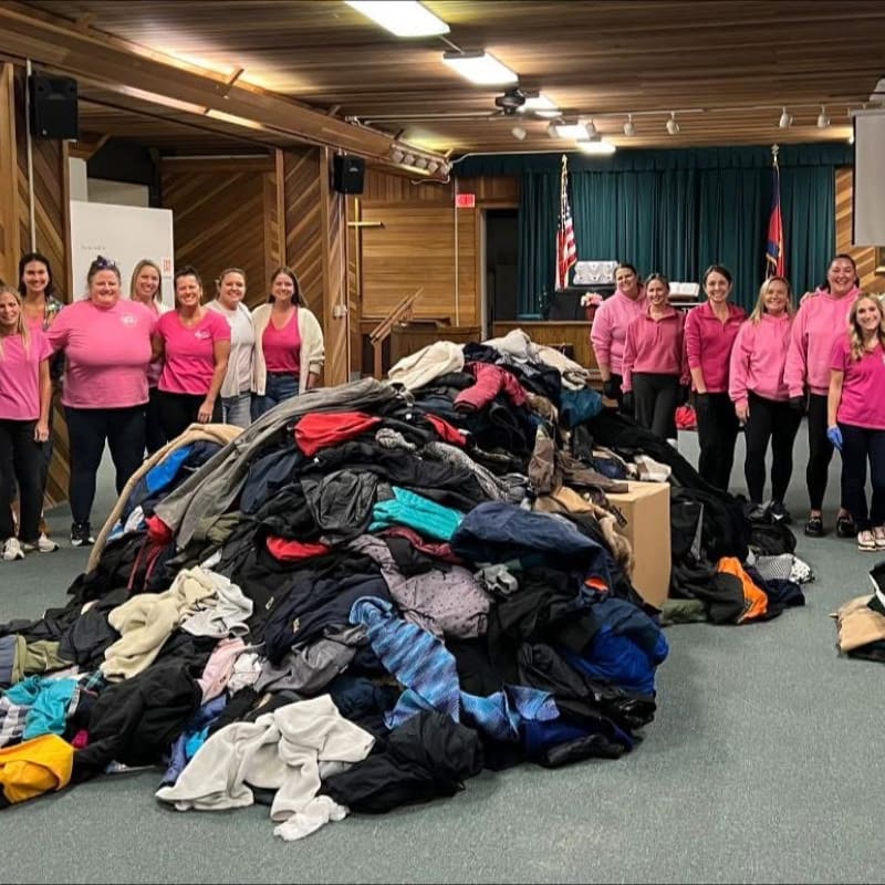 A group of women in pink shirts stand behind a large pile of donated clothing in a room with a stage and American flag in the background.