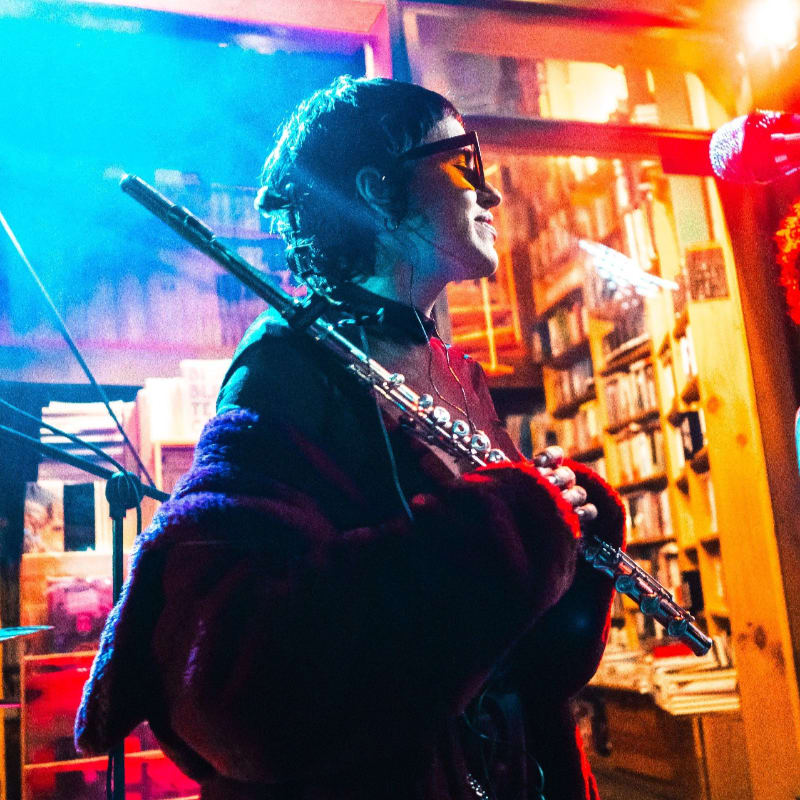 A person in the foreground plays a flute in front of a dimly lit background filled with bookshelves.