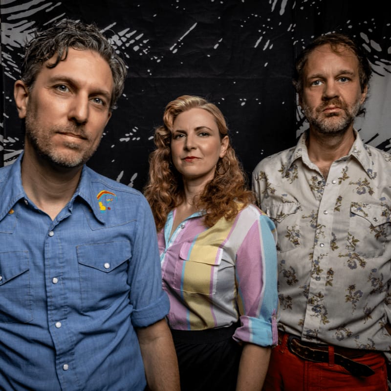 Three people stand in front of a black and white patterned backdrop, with two men on either side of a woman in the center.